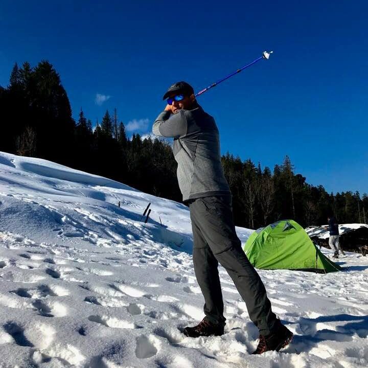 A person in winter gear swings a golf club on a snowy mountain trail, hitting a bright orange golf ball. Pine trees and distant peaks are in the background.