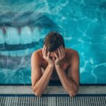 Tired swimmer sitting by the pool with a faint overlay of teeth in the background, showing how poor sleep impacts both oral health and swimming performance.